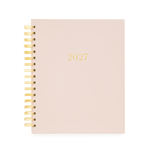 A 2027 spiral-bound planner with a pale blush pink cover and gold foil lettering, bound with a gold wire coil
