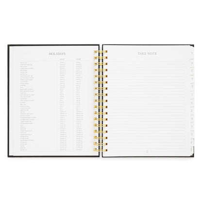 An open spiral-bound planner with a black cover, gold wire binding, and white pages. The left page is headed 'HOLIDAYS' with a list of all the holidays for 2027 and 2028, and the right page is headed 'TAKE NOTE' with lined writing space