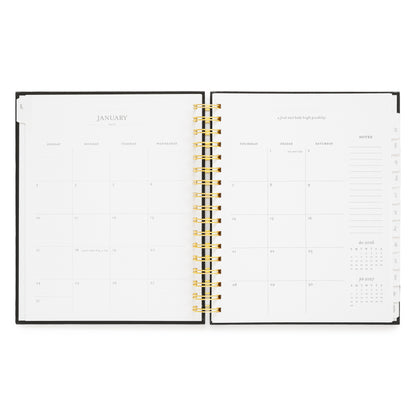 An open weekly planner with a black cover and gold spiral binding, showing the January monthly calendar page with a box for each day of the month, an inspirational quote, and notes