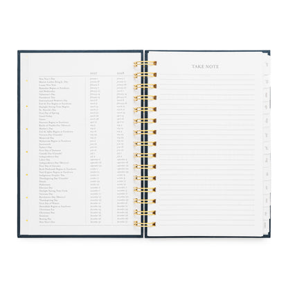 An open spiral-bound planner showing two interior pages: a left page with a dense list of holiday dates for 2027 + 2028, and a right page with a lined 'TAKE NOTE' section.