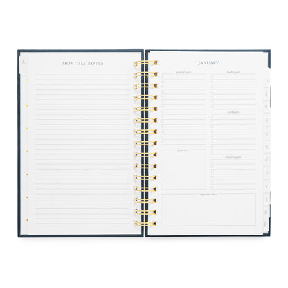 An open spiral-bound planner showing the January page with lined monthly notes on the left and task and goal setting sections on the right with a dark navy cover