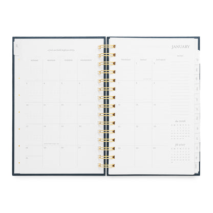 An open spiral-bound planner with a dark navy cover visible. The layout is a January 2027 calendar with spaces for each day of the month, an inspirational quote, and a notes section on the right