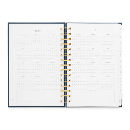 An open spiral-bound planner showing the interior pages for 2027 and 2028 with all dates of the year, with a dark navy cover and gold spiral
