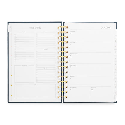 An open weekly spiral planner with interior pages visible, featuring a dark navy cover, gold spiral binding, and white pages with black text and lines. The left page shows weekly planning sections, and the right page shows a weekly January calendar layout with daily boxes.