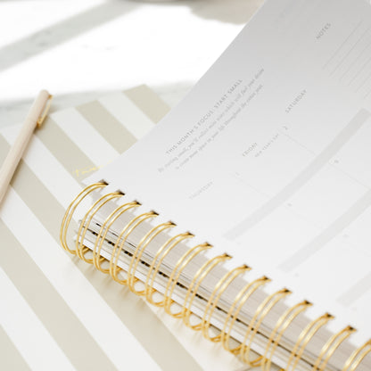 A close-up of a spiral-bound planner with gold wire rings, showing printed calendar pages with prompt 'this month's focus: start small', placed on a beige and white striped flat monthly planner