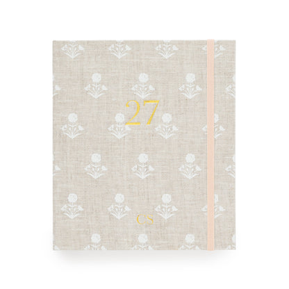 A 2027 fabric planner  with a beige linen cover, featuring a white floral pattern, a gold foil '27' on the front, and a pale pink elastic closure band with "CS' in the bottom center