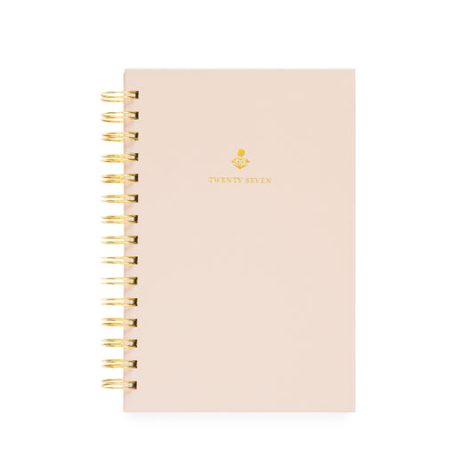 A pale blush pink spiral-bound planner with gold wire binding and a small gold floral design with 'TWENTY SEVEN' on the cover
