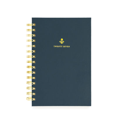 A navy spiral-bound planner with gold wire binding and a small gold floral design with 'TWENTY SEVEN' on the cover
