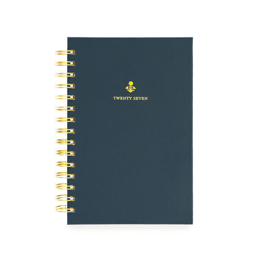 A navy spiral-bound planner with gold wire binding and a small gold floral design with 'TWENTY SEVEN' on the cover