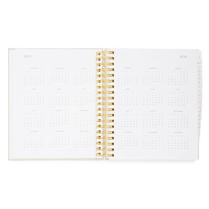 An open spiral-bound planner showing the 2027 and 2028 monthly calendar spreads on white pages, with a gold wire binding and tabs on the right showing all the calendar months