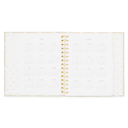An open fabric spiral-bound planner showing the year 2027 + 2028 with monthly grids on white pages with a gold spiral in the middle