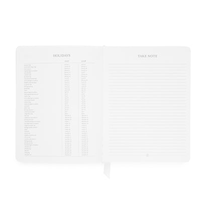 An open planner showing two interior pages: the left page has a faint 'HOLIDAYS' table with 2027 + 2028 holiday dates, and the right page has lined writing space with the heading 'TAKE NOTE'