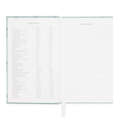 An open hardcover journal or planner showing two interior pages: a left page with a holiday list spanning 2027 and 2028, and a right page with lined space labeled TAKE NOTE with ruled lines