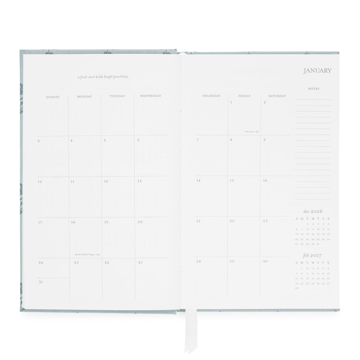 An open dusty blue agenda with blank white pages, showing a January calendar layout, a monthly grid, and lined note sections.
