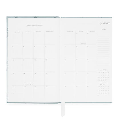 An open dusty blue agenda with blank white pages, showing a January calendar layout, a monthly grid, and lined note sections.