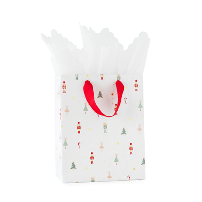 A white Christmas-themed nutcracker gift bag with red handles, decorated with small festive illustrations of nutcracker, mouse, trees, ballerina, and candy canes, filled with white tissue paper