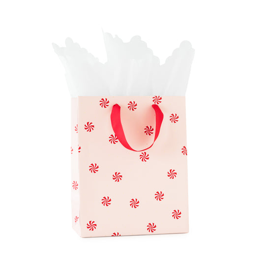 A light pink gift bag with red handles and a repeating red peppermint candy print, filled with white tissue paper