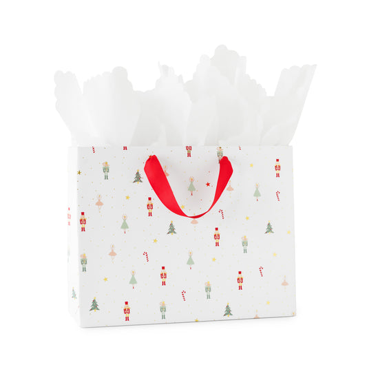 A white gift bag with a festive Christmas pattern of nutcrackers, soldiers, trees, and candy canes, featuring red ribbon handles and white tissue paper