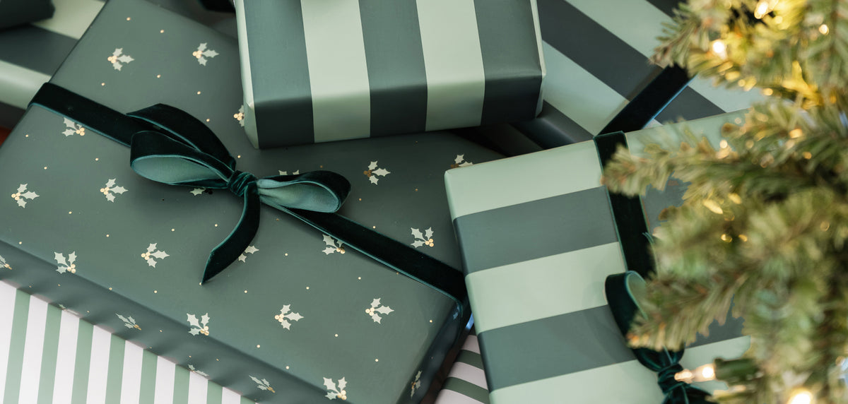A collection of wrapped Christmas presents in muted sage green and white tones, featuring striped wrapping paper, solid patterned paper with tiny white motifs, and dark green ribbon bows, styled next to a glowing Christmas tree branch.
