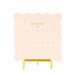 A January 2027 desk calendar on pale pink scallop edge paper with gold foil lettering, displayed on a gold metal easel stand