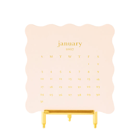 A January 2027 desk calendar on pale pink scallop edge paper with gold foil lettering, displayed on a gold metal easel stand