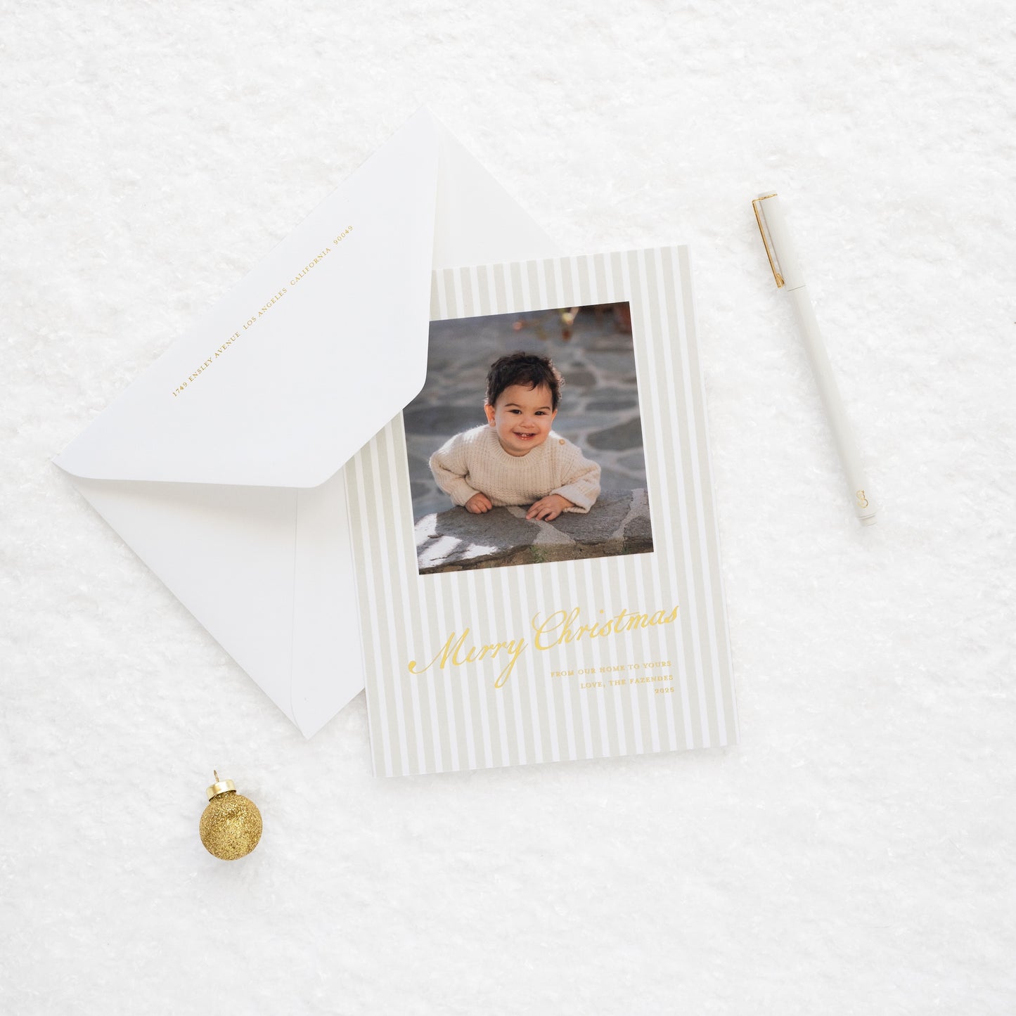 Close up of Beige stripe oversized custom holiday card with gold foil 'merry christmas from our home to yours' with photo of little boy paired with a white envelope on snow with jingle bell