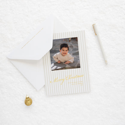 Close up of Beige stripe oversized custom holiday card with gold foil 'merry christmas from our home to yours' with photo of little boy paired with a white envelope on snow with jingle bell