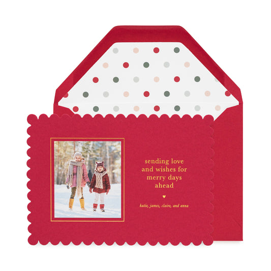Red scalloped custom holiday photo card with a photo of two kids in the snow with gold foil 'sending love and wishes for merry days ahead' paired with a red envelope with a multi dot envelope liner