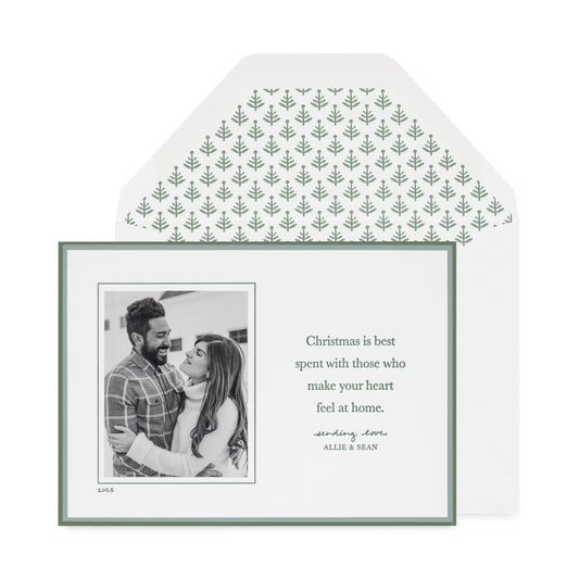 Custom holiday photo card with a black and white photo with a double green border on a white card with green letterpress ink messaging 'christmas is best spent with those that make your heart feel at home' paired with a white envelope with a green tree pattern envelope liner