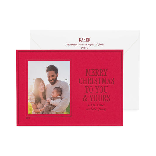 A red personalized Christmas greeting card with a photo of a family and the text 'MERRY CHRISTMAS TO YOU & YOURS' in dark red ink, shown in front of a white envelope