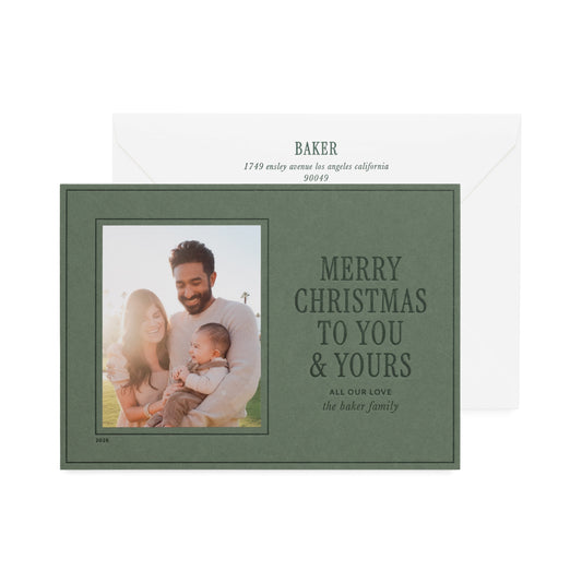 A personalized green Christmas card with a dark green border and family photo shown in front of a white envelope. The card reads 'MERRY CHRISTMAS TO YOU & YOURS' and includes the name 'BAKER' printed in dark green ink on the envelope with an address.