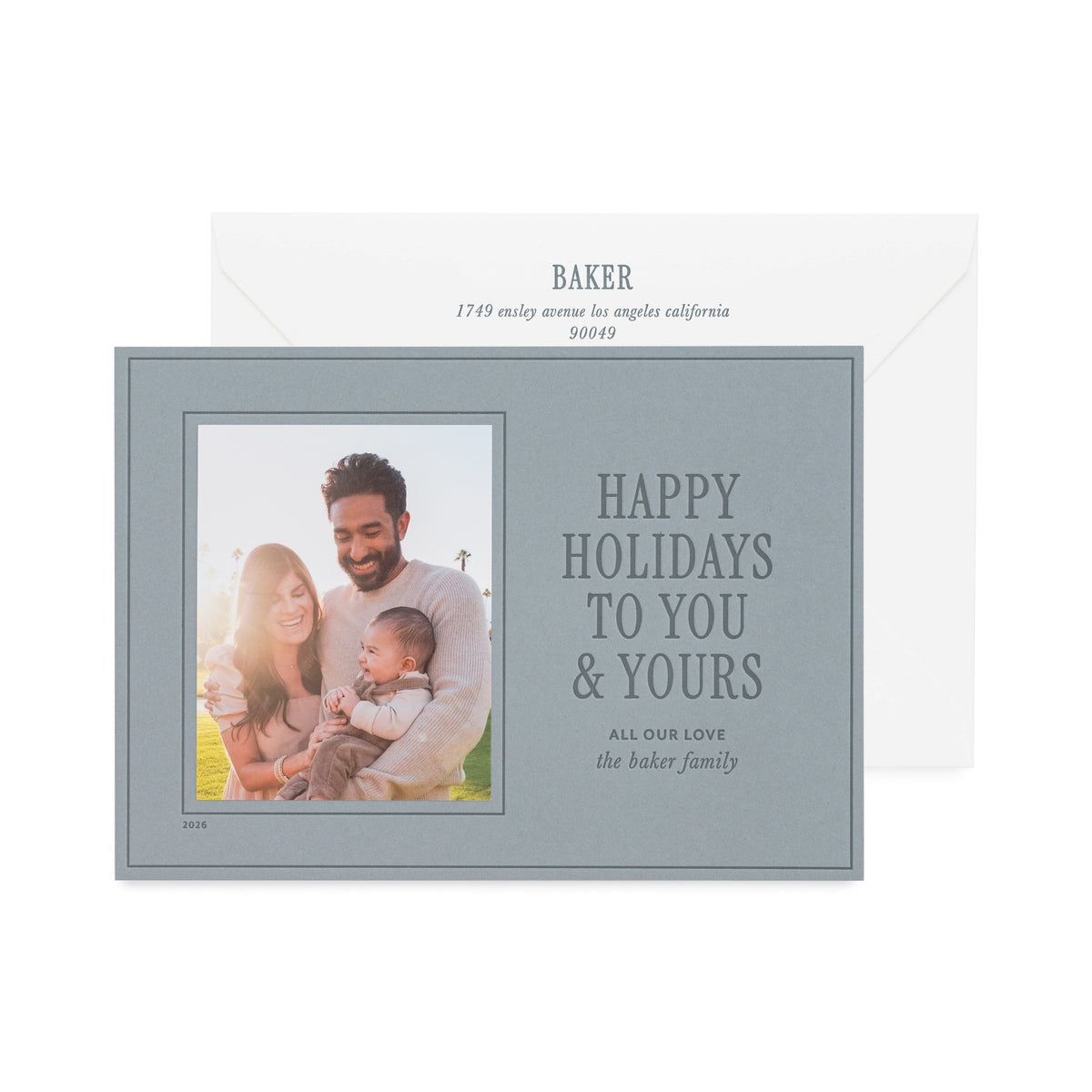 A personalized holiday card with a photo of a family and the text 'HAPPY HOLIDAYS TO YOU & YOURS' on a slate blue card with dark blue ink shown with a white envelope with 'baker" printed on it