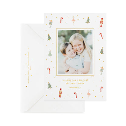 A white holiday photo card with small nutcracker, candy cane, tree, and ballerina illustrations with gold foil details and a photo of two children, printed with 'wishing you a magical christmas season,' and 'the grobeckers' paired with a white envelope