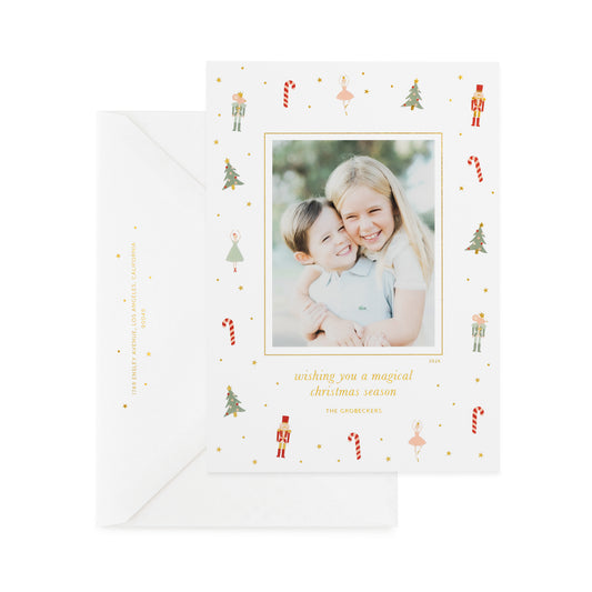 A white holiday photo card with small nutcracker, candy cane, tree, and ballerina illustrations with gold foil details and a photo of two children, printed with 'wishing you a magical christmas season,' and 'the grobeckers' paired with a white envelope
