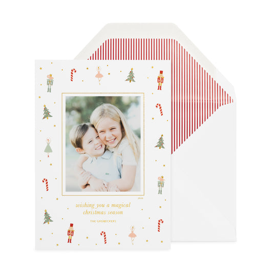 A holiday photo card decorated with small Christmas nutcracker illustrations, featuring a central photo of two children and gold text reading 'wishing you a magical Christmas season' and 'The Grobeckers.' An open white envelope with red-and-white vertical stripes sits behind the card