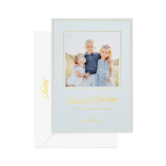 A light blue family photo greeting card with a gold foil border, gold foil script', printed with "season's greetings,' and 'from our home to yours' shown with a white envelope printed with 'bailey' 