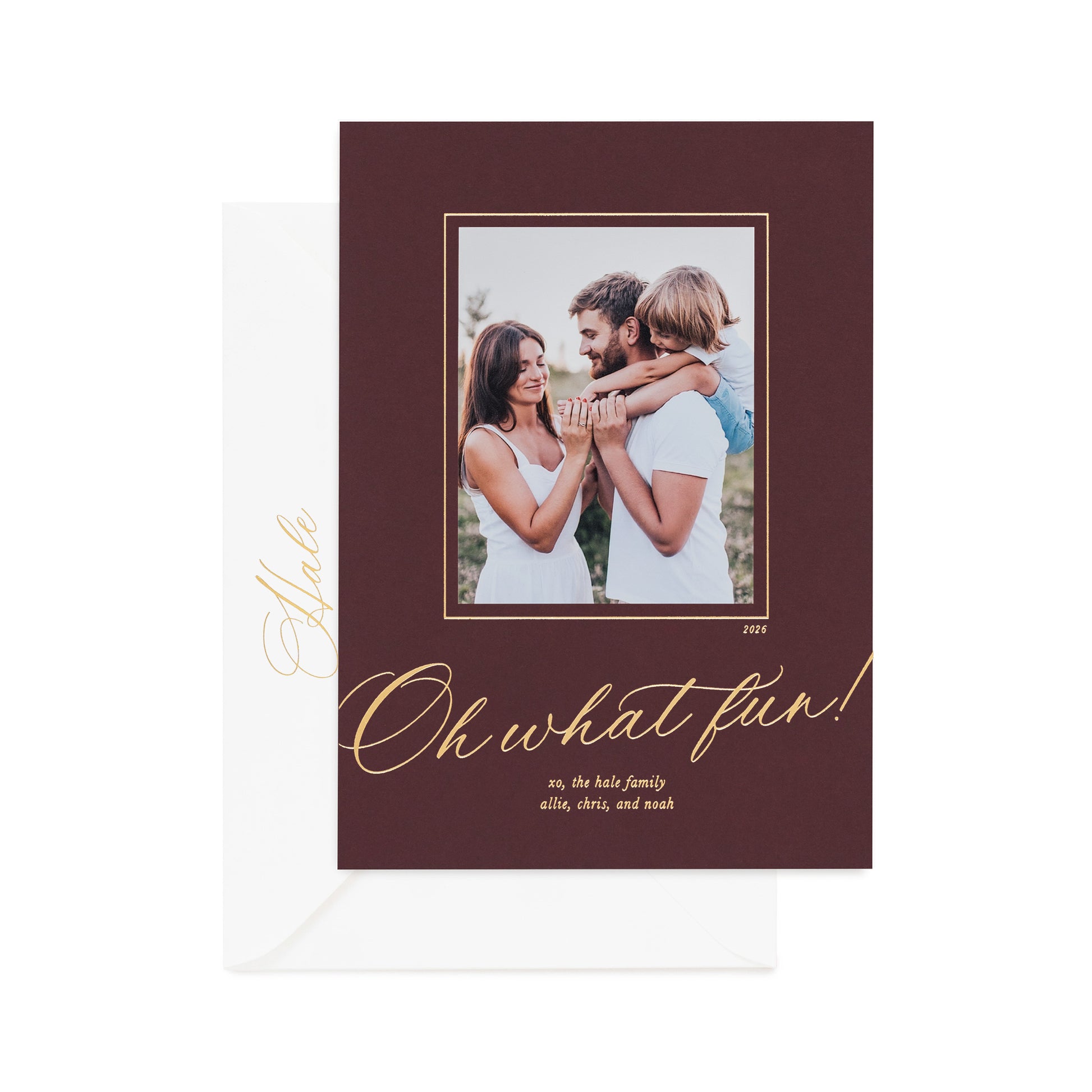 A holiday photo card on mulberry paper featuring a photo of a family, gold foil text reading 'Oh what fun!', and a white envelope behind it on a white background.