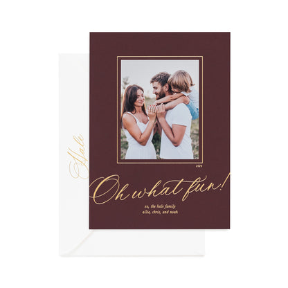A holiday photo card on mulberry paper featuring a photo of a family, gold foil text reading 'Oh what fun!', and a white envelope behind it on a white background.