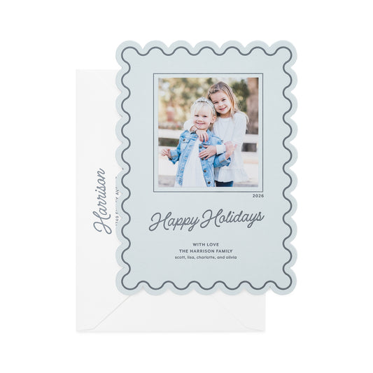 A light blue holiday photo card with a scalloped border, featuring a custom family photo of two girls, the text 'Happy Holidays' in script in dark blue ink, and the name 'Harrison' printed on the white envelope.