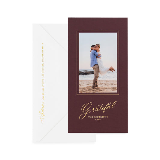 A vertical holiday card on mulberry paper, featuring a photo of a couple, gold script text and shown with a white envelope on a white background.