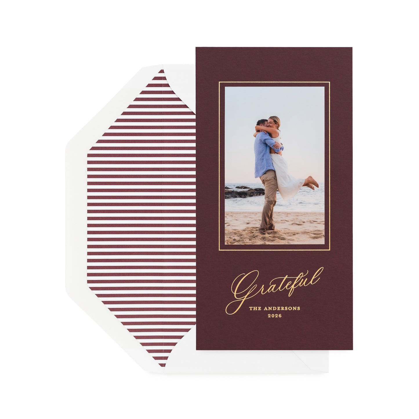 A vertical holiday card on mulberry paper with a photo of couple, gold text, shown with a white envelope featuring burgundy horizontal striped liner.