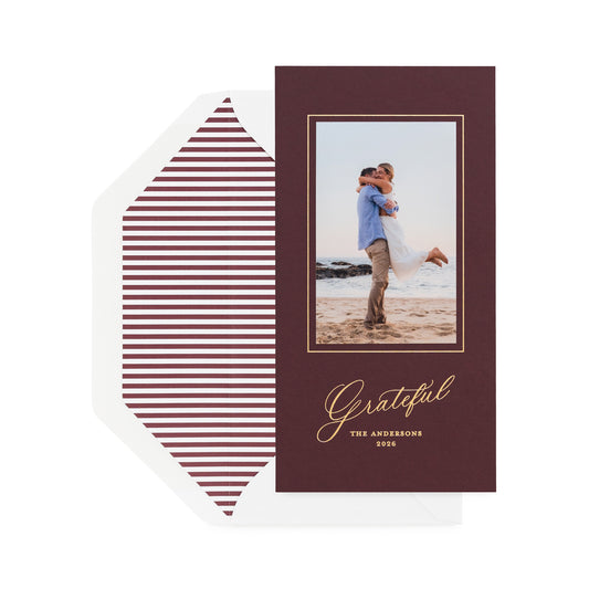 A vertical holiday card on mulberry paper with a photo of couple, gold text, shown with a white envelope featuring burgundy horizontal striped liner.