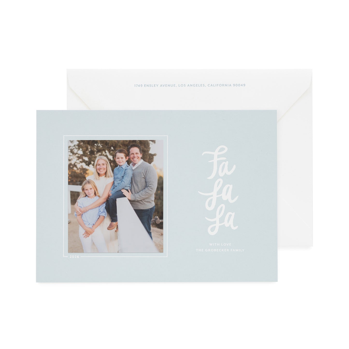 A light blue holiday greeting card with a family photo on the left and white script text reading 'Fa La La' on the right, shown in front of a white envelope with light blue text