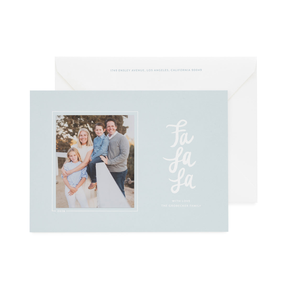 A light blue holiday greeting card with a family photo on the left and white script text reading 'Fa La La' on the right, shown in front of a white envelope with light blue text