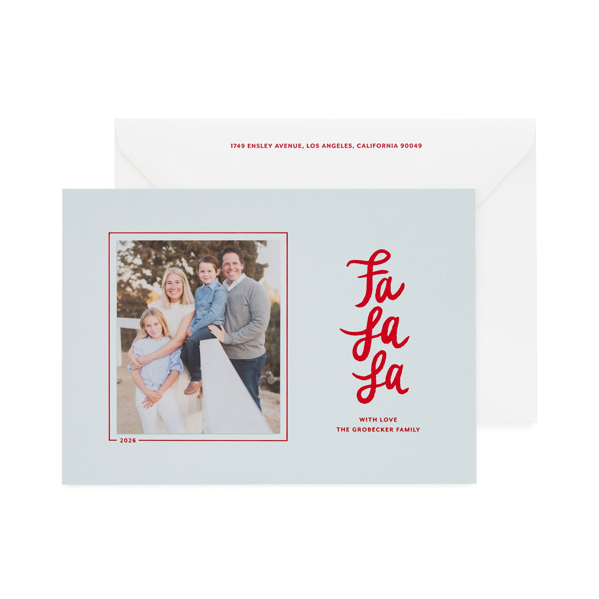 A light blue holiday greeting card with a family photo on the left and red script text reading 'Fa La La' on the right, shown in front of a white envelope with red text
