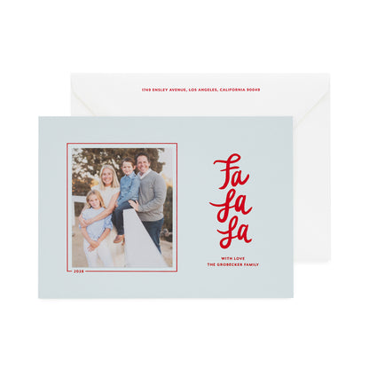 A light blue holiday greeting card with a family photo on the left and red script text reading 'Fa La La' on the right, shown in front of a white envelope with red text