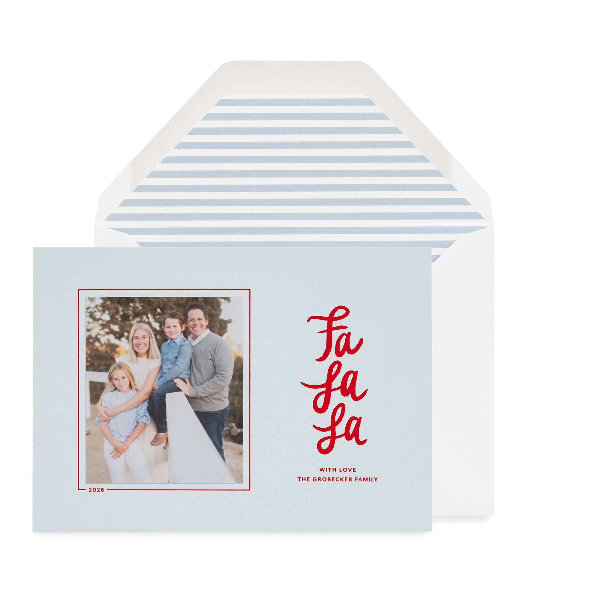 A light blue holiday greeting card with a family photo on the left and red script text reading 'Fa La La' on the right, shown in front of a white envelope with a light blue envelope liner