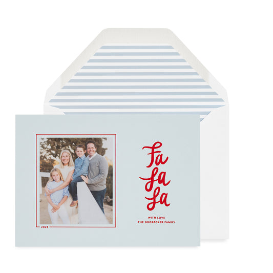 A light blue holiday greeting card with a family photo on the left and red script text reading 'Fa La La' on the right, shown in front of a white envelope with a light blue envelope liner