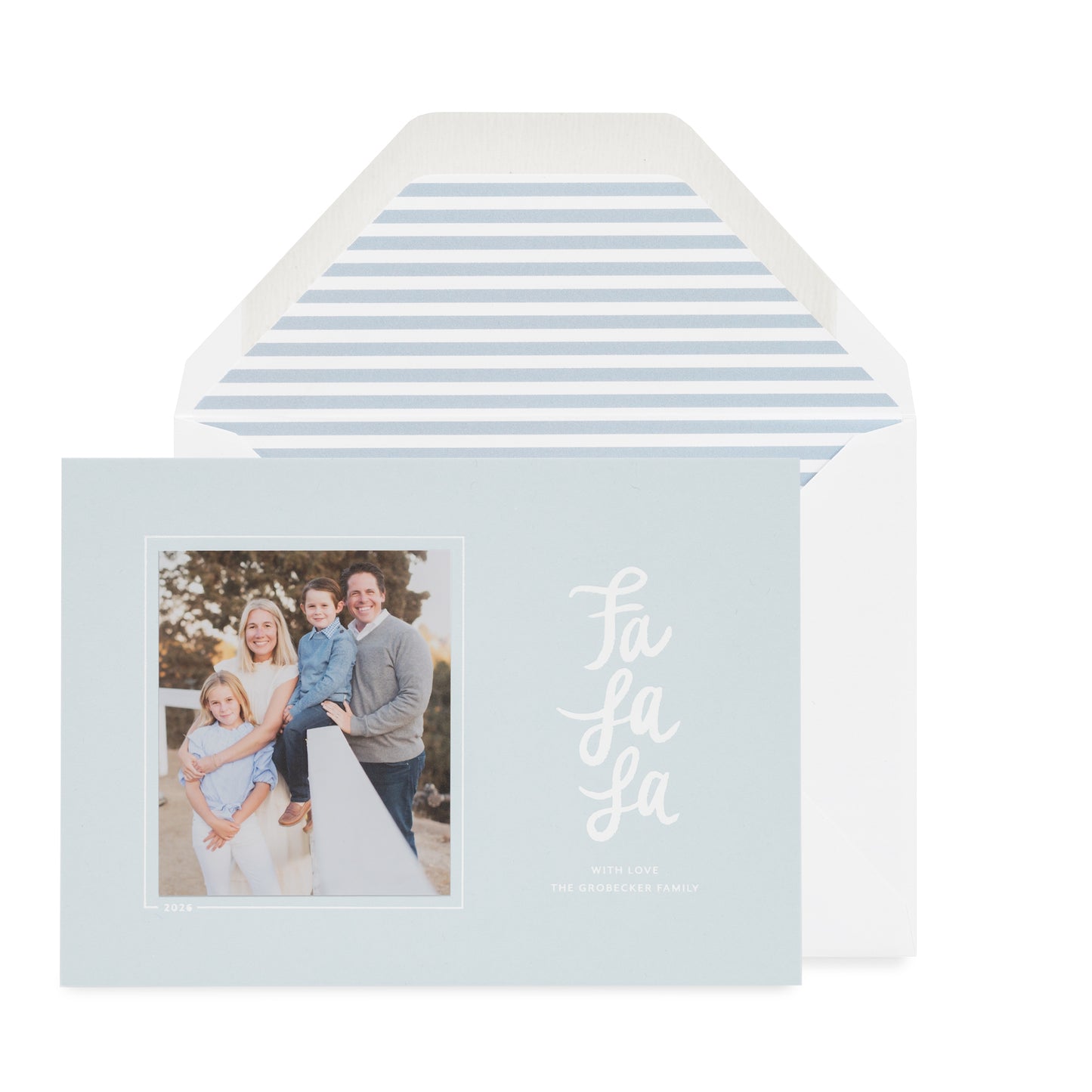 A light blue holiday greeting card with a family photo on the left and red script text reading 'Fa La La' on the right, shown in front of a white envelope with a light blue stripe envelope liner