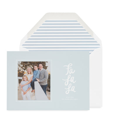 A light blue holiday greeting card with a family photo on the left and red script text reading 'Fa La La' on the right, shown in front of a white envelope with a light blue stripe envelope liner